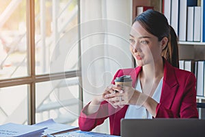 Financial, Asian businesswoman in red suit holding cup of coffee sitting on desk in office, having computer for doing accounting