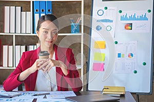 Financial, Asian businesswoman in red suit holding cup of coffee sitting on desk in office, having computer for doing accounting