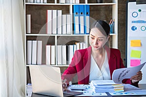 Financial, Asian businesswoman in red suit holding cup of coffee sitting on desk in office, having computer for doing accounting