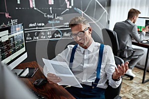 Financial matter aligned. Stylish businessman, trader looking through papers, while sitting by desk in front of computer