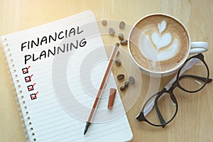 Financial Planning message and check list marks on notebook, glasses, pencil and coffee cup on wooden table with sunlight in