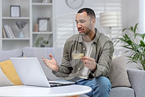 Financial problems. Upset African American young man sitting at home in front of laptop, looking worriedly at screen