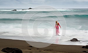 Fine art picture about a blonde, red long dress woman standing on a rock in the waving sea next to a black sand beach.
