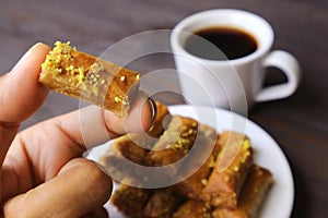 Fingers Holding a Pistachio Nuts Baklava Pastries with Blurry Turkish Coffee and a Plate of Sweets in Background