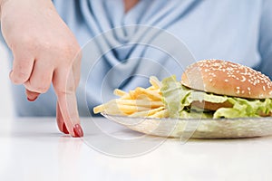 Fingers of a young lush fat woman in casual blue clothes on a white background, the choice between healthy food and fast