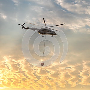 A fire fighter helicopter with a full basket of water flies against a beautiful sunset sky