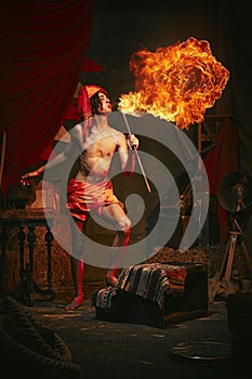 Fire show. Young man in turban breathes fire, displaying dramatic flame burst over dark retro circus backstage