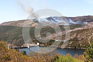 Fire with smoke on the mountains and a blue lake with a dam below