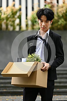 Fired Asian businessman holding cardboard box with belongings walking out of the office building. Unemployment concept