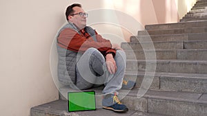 Fired caucasian male worker sitting on stairs in depression with green screen on tablet computer.