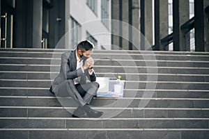 Fired male office worker in medical mask sitting on stairs in depression with box of stuff. Unemployed businessman lost