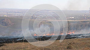 Firefighters in uniform extinguish a natural fire. Burning field on the background of the plant, the city. Dry grass in