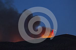 Firefighting plane soars from behind a hill dropping flame retardant. Last run of the night after sunset backlit smoke and fire