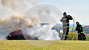 Fireman stands by a burning car and extinguishes, white smoke and flames coming out of the car