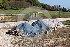 Firewood stacked in four piles covered with nylon protection against rain next to railroad tracks surrounded with grass and trees