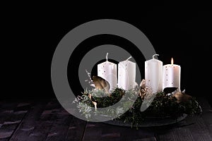 First Advent - Advent wreath from fir and evergreen branches with white burning candles on dark wooden table. Black background.