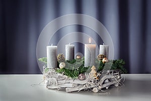 First advent with a burning candle on a wreath of white painted wood, fir branches and Christmas decoration against a purple-grey