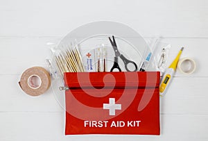 First aid kit in red bag on wooden table. Kit with patch, pills, scissors, masks, thermometer, elastic bandages. Top view,