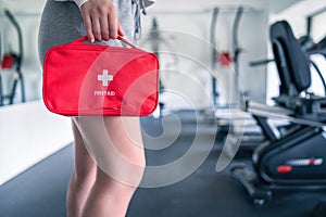 First aid kit red box in the instructor female hand in fitness gym opposite the sport equipment and jogging simulators. Healthy