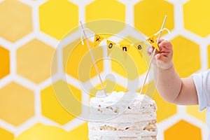 First birthday. A baby`s hand reaches for the birthday cake. Yellow background with a decor in the form of a honeycomb and