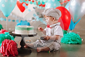 First birthday cakesmash. Adorable baby boy sitting on concrete floor and eating cake in decorated studio location