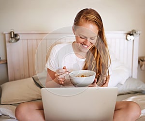 But first breakfast before I check updates. Shot of a young woman eating breakfast and using a laptop at home.