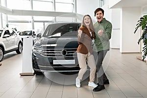 First family car. Happy young Caucasian couple shaking fists, making YES gesture after buying new auto at dealership