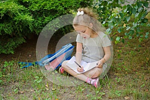 First grader sits having crossed legs under a tree and does homework.
