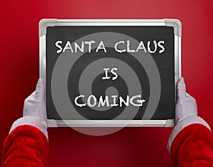 First person angle view of Santa Claus holding a black chalk board written with SANTA CLAUS IS COMING on red