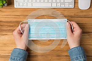 First person top view photo of female hands holding medical facemask keyboard mouse and plant on isolated wooden table background