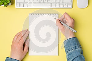 First person top view photo of female hands holding pen over open notebook plant white keyboard mouse on isolated yellow