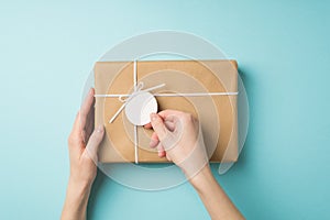 First person top view photo of girl`s hands holding pricetag on craft paper giftbox with small white bow on isolated pastel blue