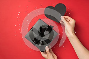 First person top view photo of hand holding black paper heart and giftbox with black ribbon bow and tag sequins on isolated red