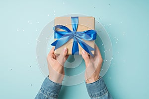 First person top view photo of hands in denim shirt holding craft paper giftbox with vivid blue ribbon bow over sequins on