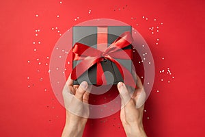 First person top view photo of hands holding black giftbox with red ribbon bow and tag shiny sequins on isolated red background