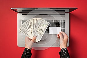 First person top view photo of hands holding fan of dollars and small rectangular white card over open laptop on  red
