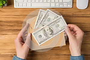 First person top view photo of hands holding open craft paper envelope with money dollars flowerpot white keyboard and mouse on