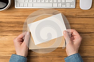 First person top view photo of hands holding open craft paper envelope and white card cup keyboard and mouse on isolated wooden