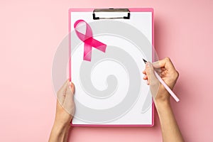 First person top view photo of hands holding pink pen and pink clipboard with paper sheet and pink ribbon symbol of breast cancer