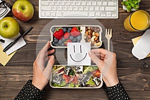 First person top view photo of hands holding sticky note with drawn smiling face over lunchboxes with healthy food apples glass of