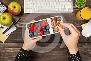 First person top view photo of woman`s hands holding lunchbox with healthy meal nuts and berries over apples glass of juice