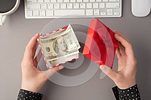 First person top view photo of woman`s hands holding open red giftbox with money hundred dollars banknotes white keyboard mouse