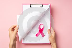 First person top view photo of woman`s hands holding pink clipboard and turning page pink ribbon symbol of breast cancer awarenes