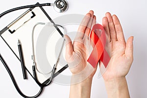 First person top view photo of woman`s hands holding red ribbon in palms over clipboard pen and stethoscope symbol of aids