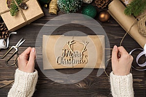 First person top view photo of woman`s hands in sweater decorating kraft paper package with merry christmas wooden text and twine