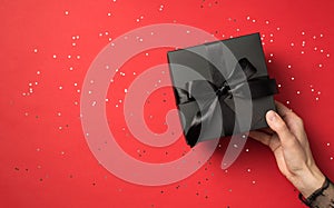First person view above photo of black giftbox with ribbon wrapped as bow in woman hands and confetti around isolated on the red