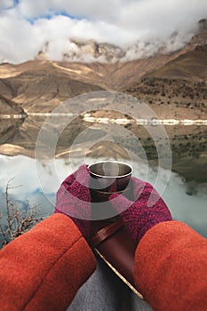 A first-person view of women`s hands in coats and mittens are holding a metal mug with tea or coffee and a leather