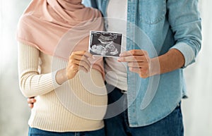 First portrait of our baby. Happy muslim pregnant woman and her husband showing sonogram picture to camera