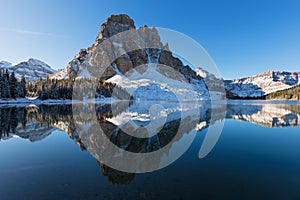 First snow in Canadian mountains. Yellow larch trees reflect like a mirror in Sunburst Lake below a rocky peak