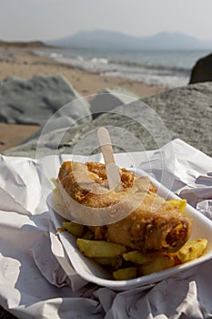 Fish and chips, battered cod, in a tray wrapped in paper with a wooden fork on a beach. Traditional British food